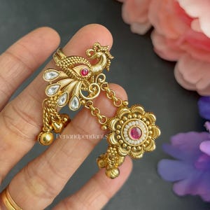 May include: A gold-toned ring featuring a peacock motif with a small red stone, white teardrop-shaped accents, and a dangling floral charm with a central red stone and white beads. The text Penandpendants is visible on the metal surface.