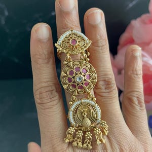 May include: A gold-toned multi-segment ring featuring three distinct circular and almond-shaped sections. The design includes small white beads, embedded pink stones, and a central peacock motif at the base with dangling gold-toned charms.