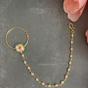 May include: A gold-toned nose ring featuring a circular hoop and a central floral-shaped ornament with a pink stone, green enamel detailing, and white accents. A delicate gold chain adorned with small white beads connects to a hook clasp.