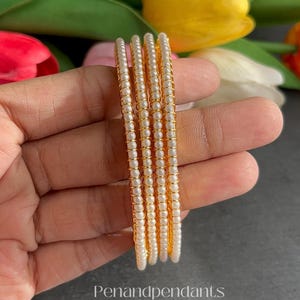 May include: A set of five gold-toned bangles, each adorned with small, white, pearl-like beads. The bangles are held in a hand, with a blurred background of colorful tulips. The text "Penandpendants" is visible.