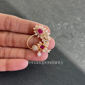 May include: A gold-colored nose ring with a floral design. The ring features a large, oval, ruby-colored stone surrounded by small, white pearls and clear crystals. The text "Penandpendants" is visible.