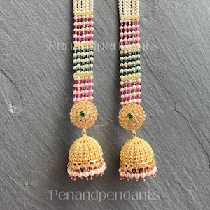 May include: A pair of gold-colored earrings with a pearl and bead design. The earrings feature a gold-colored bell-shaped bottom with a pearl trim and a green gem in the center. The earrings are made of multiple strands of white pearls, with sections of pink and green beads.