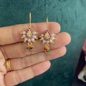 May include: A pair of gold drop earrings featuring a central teardrop-shaped gold plate surrounded by small pink stones and white pearls. Each earring has a dangling charm with a small red stone and a small green stone at the bottom.