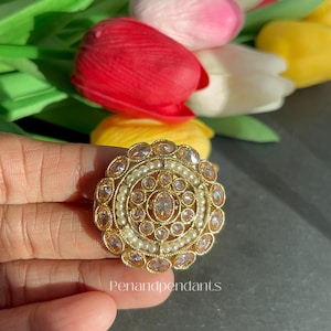 May include: A gold-toned ring with a circular design, featuring clear gemstones and small pearls. The ring is held in front of a blurred background of colorful tulips. The text "Penandpendants" is visible on the ring.