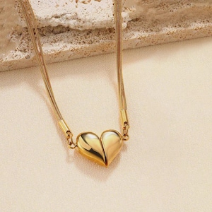 May include: A gold-colored necklace featuring a heart-shaped pendant. The pendant is split into two halves, creating a unique design. The necklace has a delicate chain and is displayed on a neutral background, highlighting the jewelry's details.