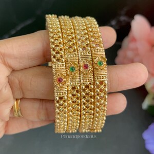 May include: A set of four gold-toned metal bangles featuring a textured beaded surface and small floral-shaped accents. Each floral accent is set with a single round red or green stone. The text Penandpendants is printed at the bottom center.