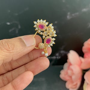 May include: A gold-toned nose ring featuring two teardrop-shaped pink stones, clear accent stones, and a border of small white pearls. The jewelry piece has a curved wire frame and includes two dangling pearl charms at the bottom base.