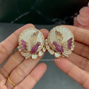 May include: A pair of round stud earrings featuring a white shell background with a gold-toned bird motif encrusted with pink and clear stones. The bird design is accented by additional clear stones along the bottom edge. The text Penandpendants is visible.