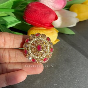 May include: A gold-toned ring with a central oval ruby surrounded by smaller red gems and pearls. The ring has a circular design with the text "Penandpendant" visible.