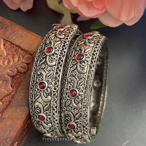 May include: Two silver-toned metal bangles featuring intricate embossed floral patterns and small, round, deep red stones set at regular intervals. The bangles have a textured, oxidized finish and a wide, cylindrical band design with visible metalwork.