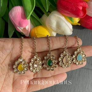 May include: Close-up of five gold-toned, floral-shaped hair accessories with various colored center stones: yellow, clear, green, pink, and blue. Each piece is connected to a chain. The text "Penandpendants" is visible.
