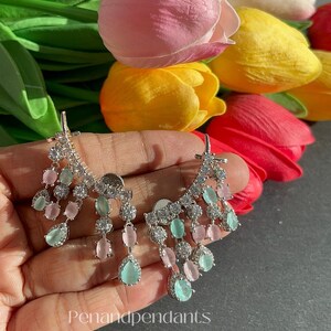 May include: Silver-toned chandelier earrings with pink and teal teardrop-shaped and oval-shaped stones. The earrings are set against a backdrop of colorful tulips. The text "Penandpendants" is visible.