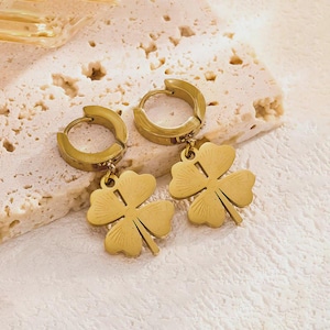 May include: A pair of gold-toned hoop earrings featuring dangling four-leaf clover charms. Each clover charm has a subtle etched sunburst pattern on its surface. The earrings are constructed with a hinged hoop mechanism for secure closure.