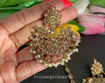 May include: Gold-toned chandelier earrings with clear and pink gemstones and dangling pearls. The earrings have an ornate floral design and are held in a hand. The text "Penandpendant" is visible.