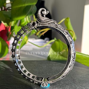 May include: A silver-toned bangle bracelet with a peacock design. The bracelet features intricate detailing, including a peacock figure at the top and decorative patterns along the band. A small turquoise stone accents the clasp. The text "Penandpendant" is visible.
