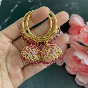 May include: Gold hoop earrings featuring bell-shaped jhumka pendants adorned with pink teardrop-shaped stones and small red beads. The metalwork includes intricate filigree patterns and the text Penandpendants is visible on the jewelry.