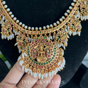 Puede incluir: Collar dorado con detalles intrincados, con un colgante central con una figura de deidad. El collar está adornado con detalles rojos, verdes y blancos, y pequeñas perlas blancas. El texto "Penandpendants" es visible.