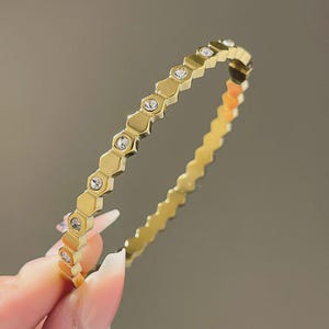 May include: A gold-toned bangle bracelet featuring a repeating hexagonal link pattern. Every other link is set with a small, clear, round faceted crystal. The metal has a polished finish, creating a continuous geometric band design.