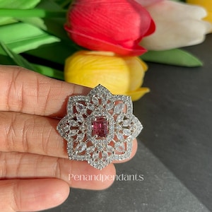 May include: A silver-toned ring with a central rectangular pink gemstone, surrounded by clear, sparkling stones in a floral design. The ring is set against a backdrop of colorful tulips. The text "Penandpendant" is visible.