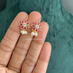 May include: A pair of gold-tone earrings featuring a flower-shaped design with marquise-cut red stones surrounding a central clear round stone. Two small white pearls hang from the base of each flower. The text Penandpendants is visible.