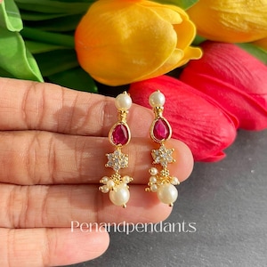 May include: Gold-toned earrings with a teardrop-shaped red gemstone, a starburst of small clear stones, and a cluster of pearls. The earrings are displayed against a backdrop of colorful tulips. The text "Penandpendants" is visible.