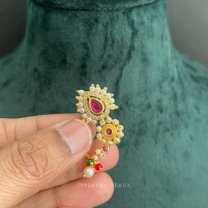 May include: A gold-toned nose ring featuring a teardrop-shaped pink stone, a circular pink stone, and small white pearls. The jewelry piece includes a small green bead and a red bead at the bottom, with the text Penandpendants visible on the surface.