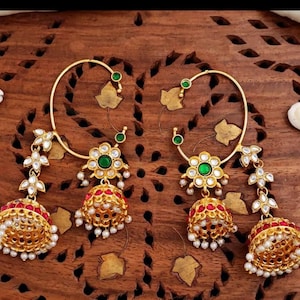May include: A pair of gold-toned earrings with intricate detailing. Each earring features a large hoop with green and white accents, a floral centerpiece, and a dangling bell-shaped charm with red and white embellishments. The earrings are displayed on a wooden surface.
