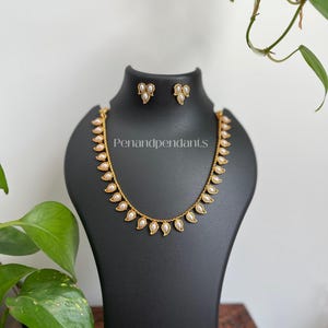 May include: A gold-toned necklace and matching earrings set displayed on a black bust. The necklace features a delicate chain with teardrop-shaped pendants, each holding a small, white pearl. The earrings mirror the pendant design. The text "Penandpendant" is visible.