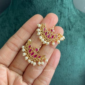 May include: A pair of gold-toned crescent moon earrings featuring marquise-cut fuchsia stones, a small star accent, and a dangling fringe of small white pearls. The earrings are crafted with a delicate metal frame and include the text Penandpendants.