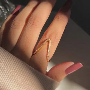 May include: A gold-colored ring in a V-shape, encrusted with small, sparkling stones. The ring is worn on a finger, with a light-colored sleeve in the background. The ring's design is modern and minimalist, suitable for everyday wear.