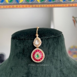 May include: A gold-toned maang tikka featuring a teardrop-shaped pendant with a central green stone surrounded by red enamel and clear crystals. A smaller teardrop-shaped accent with white beads and a clear stone sits above. Text reads Penandpendants.