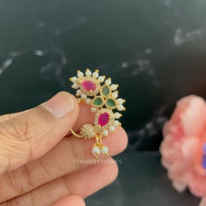 May include: A gold-tone nose ring featuring a crescent-shaped design adorned with two teardrop-shaped pink stones, two teardrop-shaped green stones, small clear crystals, and white pearls. The piece includes the text Penandpendants.