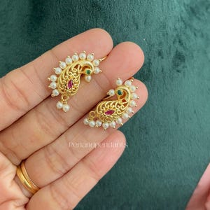 May include: A pair of gold-toned earrings featuring a paisley-shaped design adorned with small white pearls along the outer edge, a central marquise-cut pink stone, and a small green stone accent near the top, with intricate filigree metalwork.