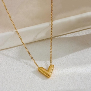 May include: A gold-colored necklace with a V-shaped pendant. The pendant and chain are made of a shiny, metallic material. The necklace is displayed on a white surface.