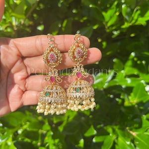 May include: Gold-toned earrings with intricate detailing, featuring red and green gemstones, and small white pearls. The earrings have a floral design and a bell-shaped bottom. The background is a blurred view of green foliage.