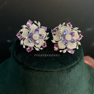 May include: A pair of flower-shaped earrings featuring central purple oval stones surrounded by pavé-set clear crystals and outer pink oval stones. The metal setting is silver-toned. The text Penandpendants is visible between the two earrings.