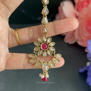 May include: A gold-toned maang tikka featuring a vertical chain of teardrop-shaped clear stones leading to a floral pendant. The pendant has a central round red stone surrounded by clear stones, a crescent base, and a dangling red bead with small white pearls.