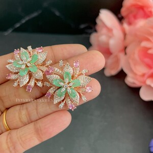 May include: A pair of rose gold-toned flower-shaped earrings featuring central marquise-cut green stones, surrounded by clear pavé-set crystals and small pink oval stones. The text Penandpendants is visible on the lower left side of the jewelry.