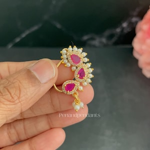 May include: A gold-toned nose ring featuring three teardrop-shaped pink stones surrounded by small white beads and clear crystals. The piece includes a dangling charm with three small white beads and the text Penandpendants printed below.