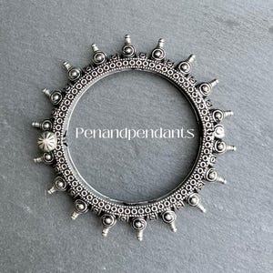 May include: A silver-colored bangle with an intricate design. The bangle features a series of small, raised circular elements and conical spikes around the outer edge. The text "Penandpendants" is visible in the center of the bangle.