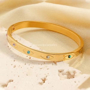 May include: A gold-colored bangle bracelet with an eye design. The bracelet features small, colorful gemstones in pink, blue, and green. The bracelet is on a textured, off-white surface.
