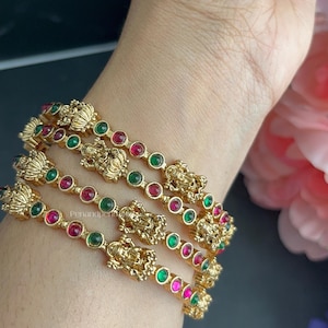 May include: A set of three gold-toned bangles featuring alternating round red and green stones. Each bangle is accented with a central embossed deity figure and floral motifs, creating a detailed metallic design worn on the wrist.