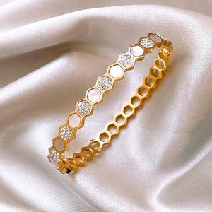 May include: Gold-toned bracelet with a honeycomb design. The bracelet features alternating sections of white, iridescent material and sparkling, clear crystals. The bracelet is set against a white, satin-like background.