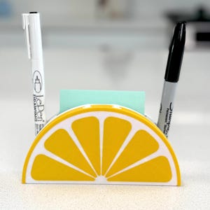 Pode incluir: Um organizador de secretária em forma de fatia de limão amarelo e branco, com uma caneta branca, um marcador preto e uma pilha de notas adesivas azuis claras. O organizador está numa superfície branca.