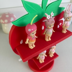 May include: A red, strawberry-shaped shelf with green leaves, holding several small figurines. The figurines are wearing hats and outfits with floral and fruit designs. A mushroom-shaped decoration is in the background.