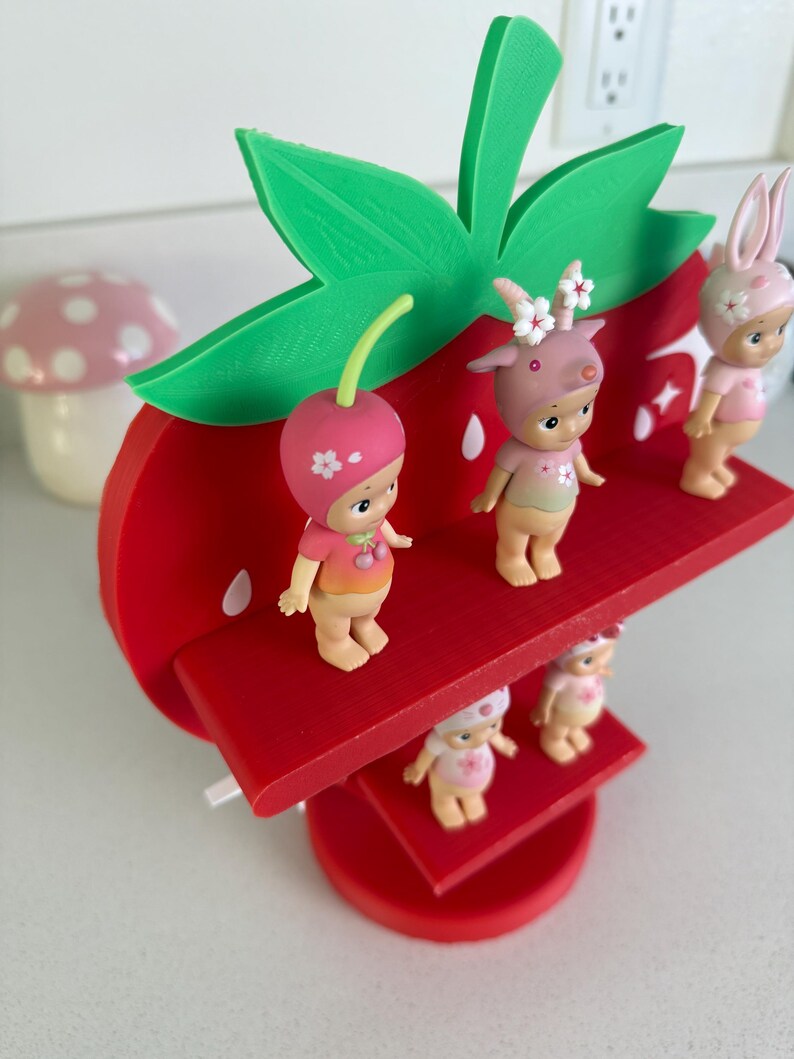 May include: A red, strawberry-shaped shelf with green leaves, holding several small figurines. The figurines are wearing hats and outfits with floral and fruit designs. A mushroom-shaped decoration is in the background.