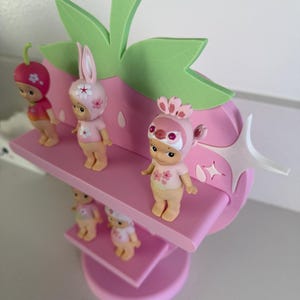 May include: A pink strawberry-shaped shelf with green leaves and white star accents, holding several small figurines. The figurines are wearing strawberry, bunny, and bird-themed hats and outfits. The shelf has multiple levels.