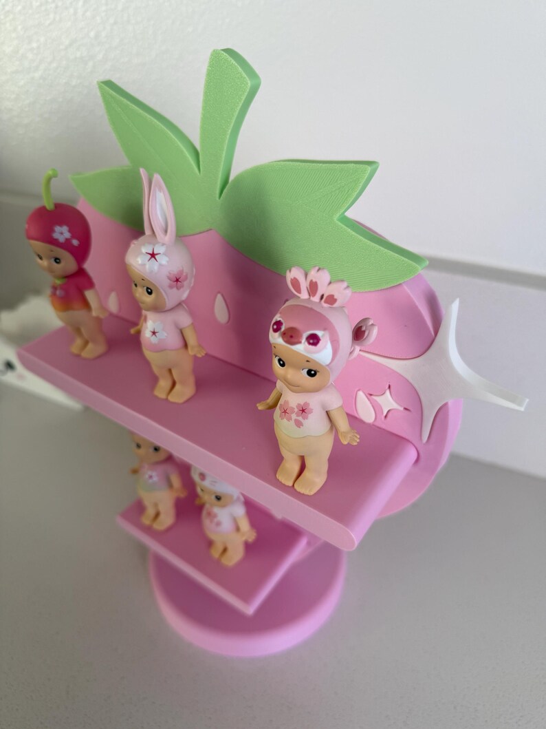 May include: A pink strawberry-shaped shelf with green leaves and white star accents, holding several small figurines. The figurines are wearing strawberry, bunny, and bird-themed hats and outfits. The shelf has multiple levels.