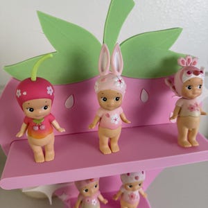 May include: A pink strawberry-shaped display stand holding six small figurines. The figurines are wearing hats and outfits with floral and fruit designs. The stand has green leaf accents and a pink shelf.