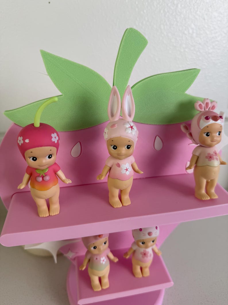 May include: A pink strawberry-shaped display stand holding six small figurines. The figurines are wearing hats and outfits with floral and fruit designs. The stand has green leaf accents and a pink shelf.